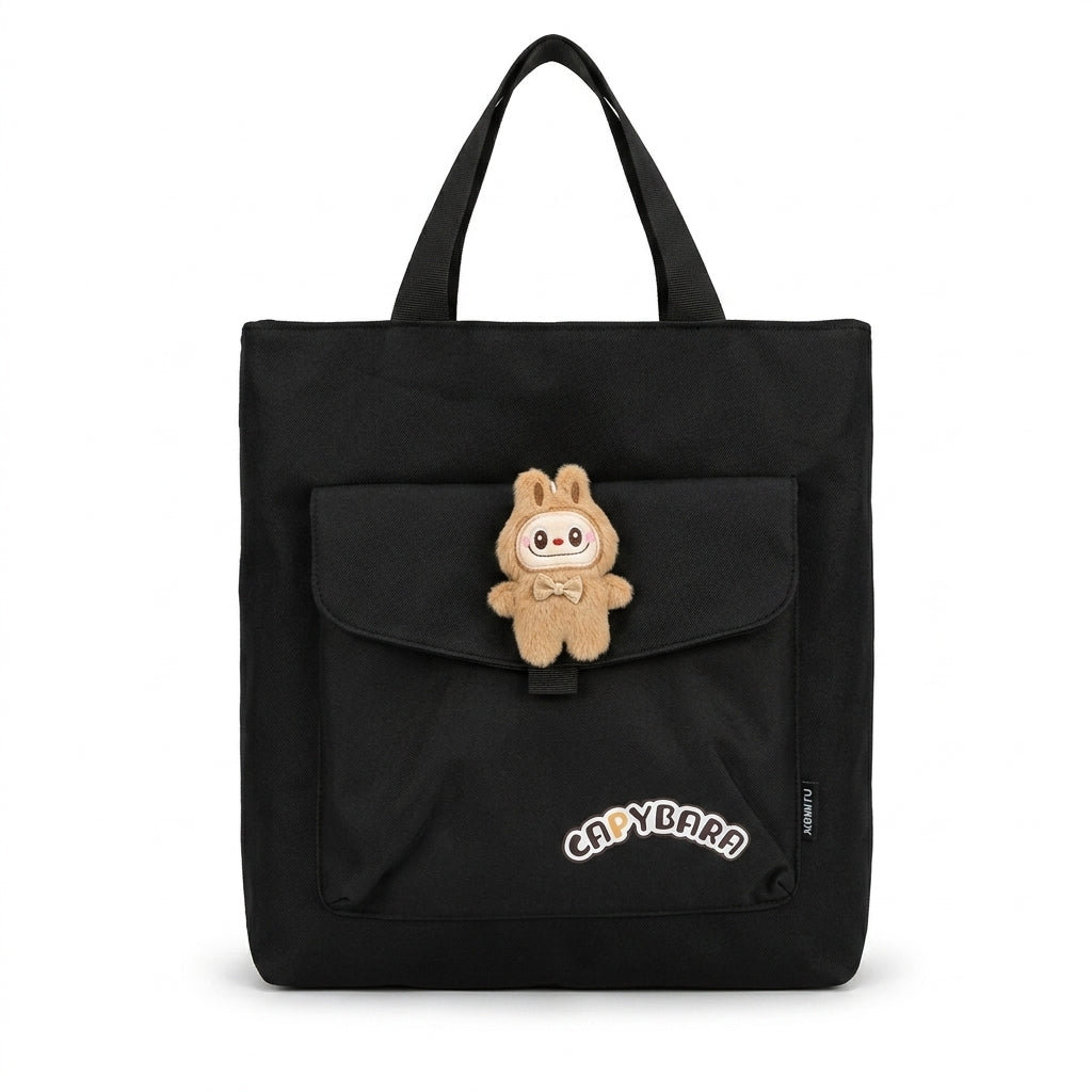 Black canvas tote bag front view featuring front flap pocket with plush character charm and Capybara text patch