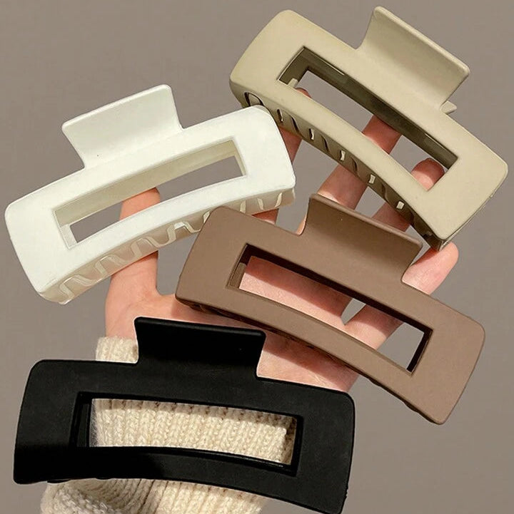 A pack of four womens matte rectangle hair claw clips in beige, white, brown, and black colors being displayed on a hand