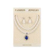 Women's white faux pearl necklace with blue teardrop pendant and matching drop earrings set on display card